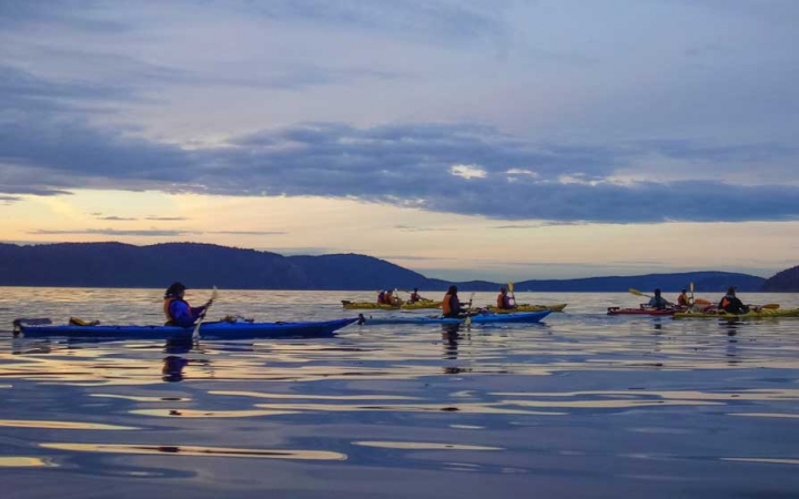 A group of students paddle kayaks on calm water in soft light. There are mountains in the background.